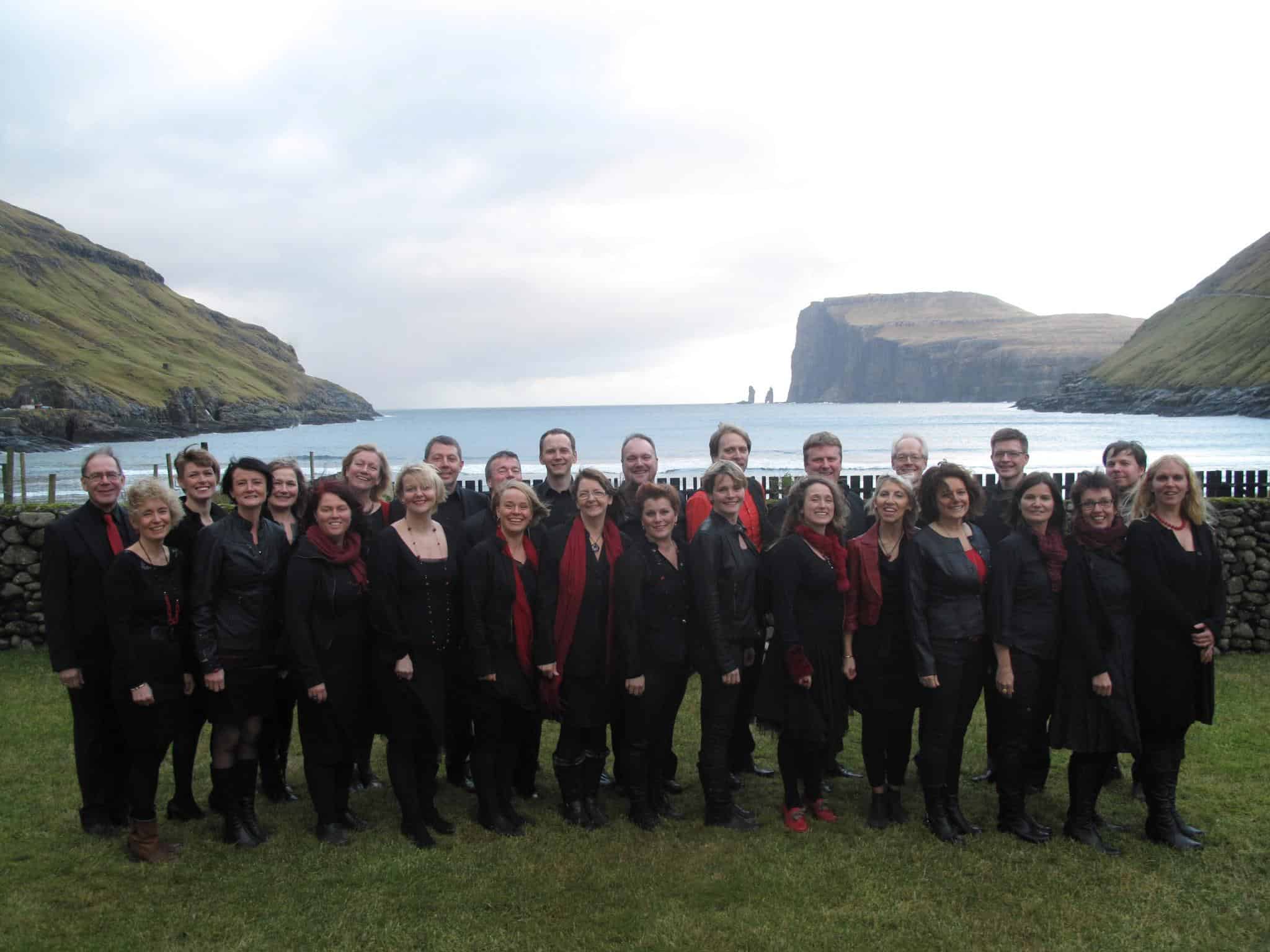 Groupe de choristes habillés en noir avec touches de rouge devant un paysage côtier spectaculaire, en Écosse.